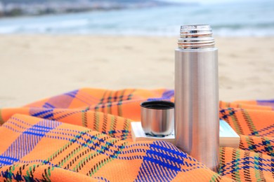 Metallic thermos with hot drink, open book and plaid on sandy beach near sea, space for text