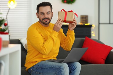 Celebrating Christmas online with exchanged by mail presents. Man with gift box during video call on laptop at home