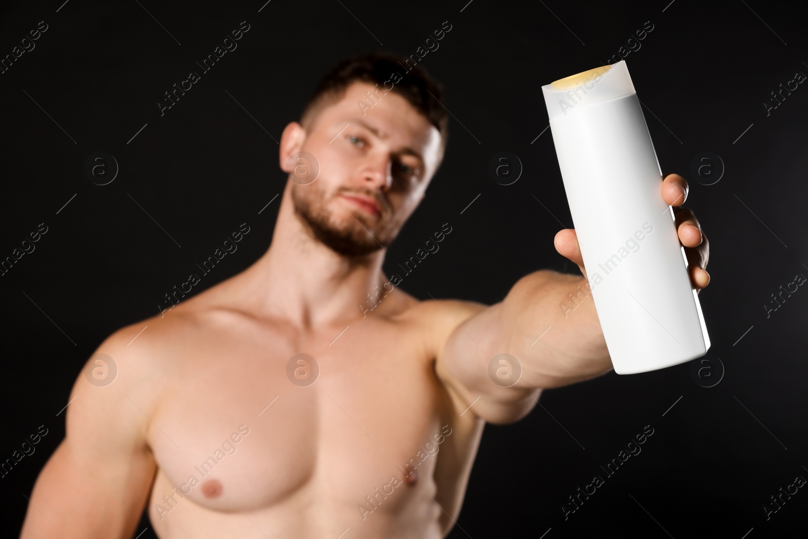 Photo of Shirtless young man holding bottle of shampoo against black background, focus on hand