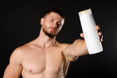 Shirtless young man holding bottle of shampoo against black background, focus on hand