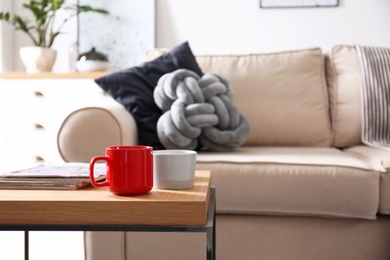 Wooden table with cups and magazines near comfortable sofa in living room interior. Space for text