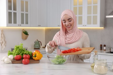 Photo of Muslim woman making delicious salad with vegetables at white table in kitchen