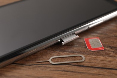 SIM card, mobile phone and ejector tool on wooden table, closeup