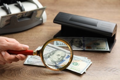 Woman checking dollar banknotes with currency detector and magnifying glass at wooden table, closeup. Money examination device