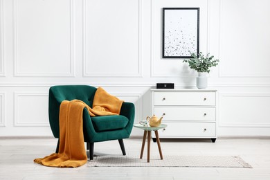 Photo of Comfortable armchair with blanket, side table and chest of drawers in room