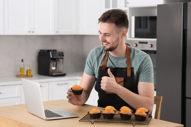 Photo of Man holding muffin near laptop and showing thumb up at table in kitchen. Time for hobby