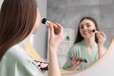 Beautiful young woman applying blusher with brush near mirror indoors