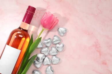 Delicious heart shaped chocolate candies, beautiful tulip and bottle of wine on pink table, flat lay. Space for text