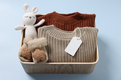 Photo of Different baby accessories, knitted sweaters and blank card in box on light blue background