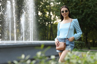 Photo of Beautiful young woman with stylish bag in sunglasses at park outdoors, space for text