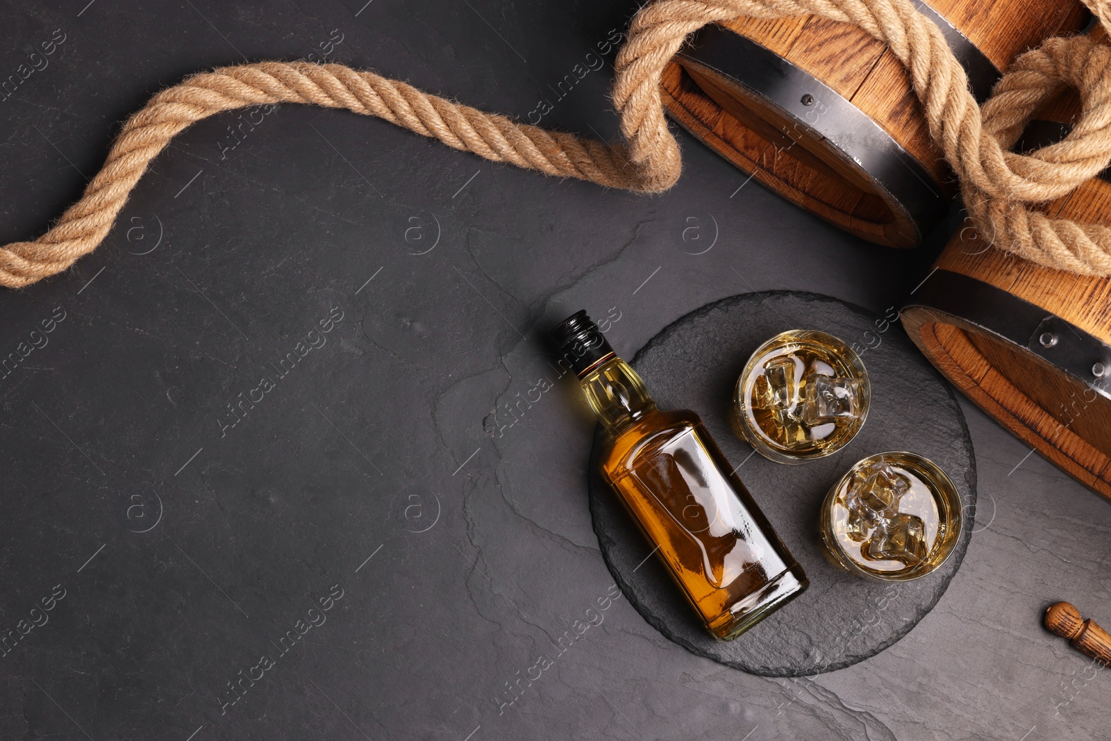 Photo of Whiskey with ice cubes in glasses, bottle, wooden barrels and rope on black table, top view. Space for text