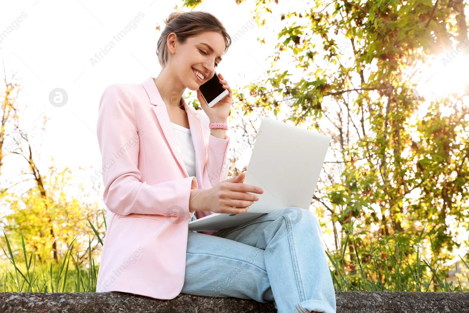 Image of Happy young woman talking on phone while using laptop outdoors