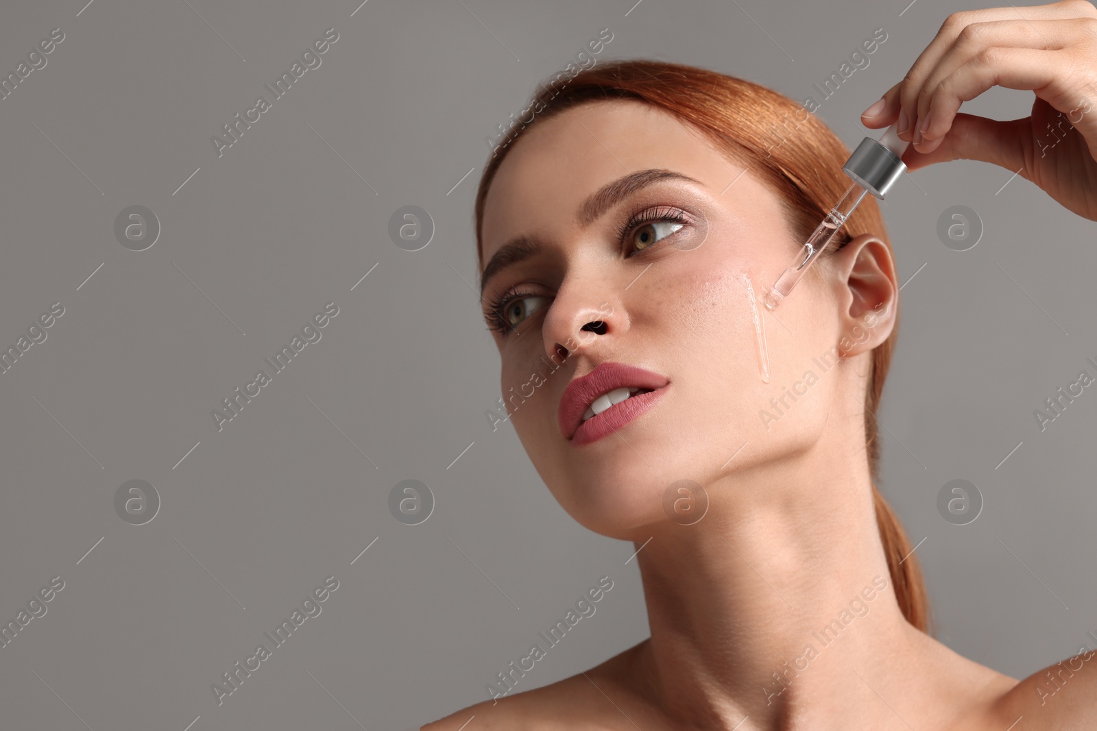 Photo of Beautiful young woman applying cosmetic serum onto her face on grey background, space for text