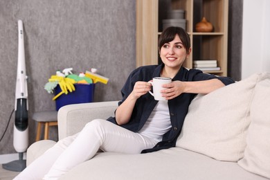 Beautiful young housewife with cup of drink resting after cleaning on sofa at home