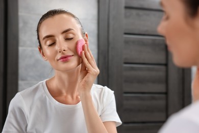 Photo of Young woman washing her face with sponge near mirror in bathroom