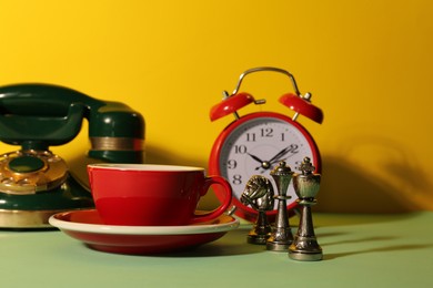 Red cup with saucer, alarm clock, vintage telephone and chessmen on light green table against yellow background