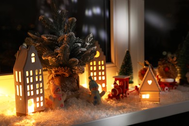 Photo of Christmas atmosphere. Beautiful glowing houses, fir trees, artificial snow and toys on window sill indoors