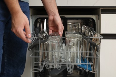 Man loading dishwasher with glass and cutlery indoors, closeup