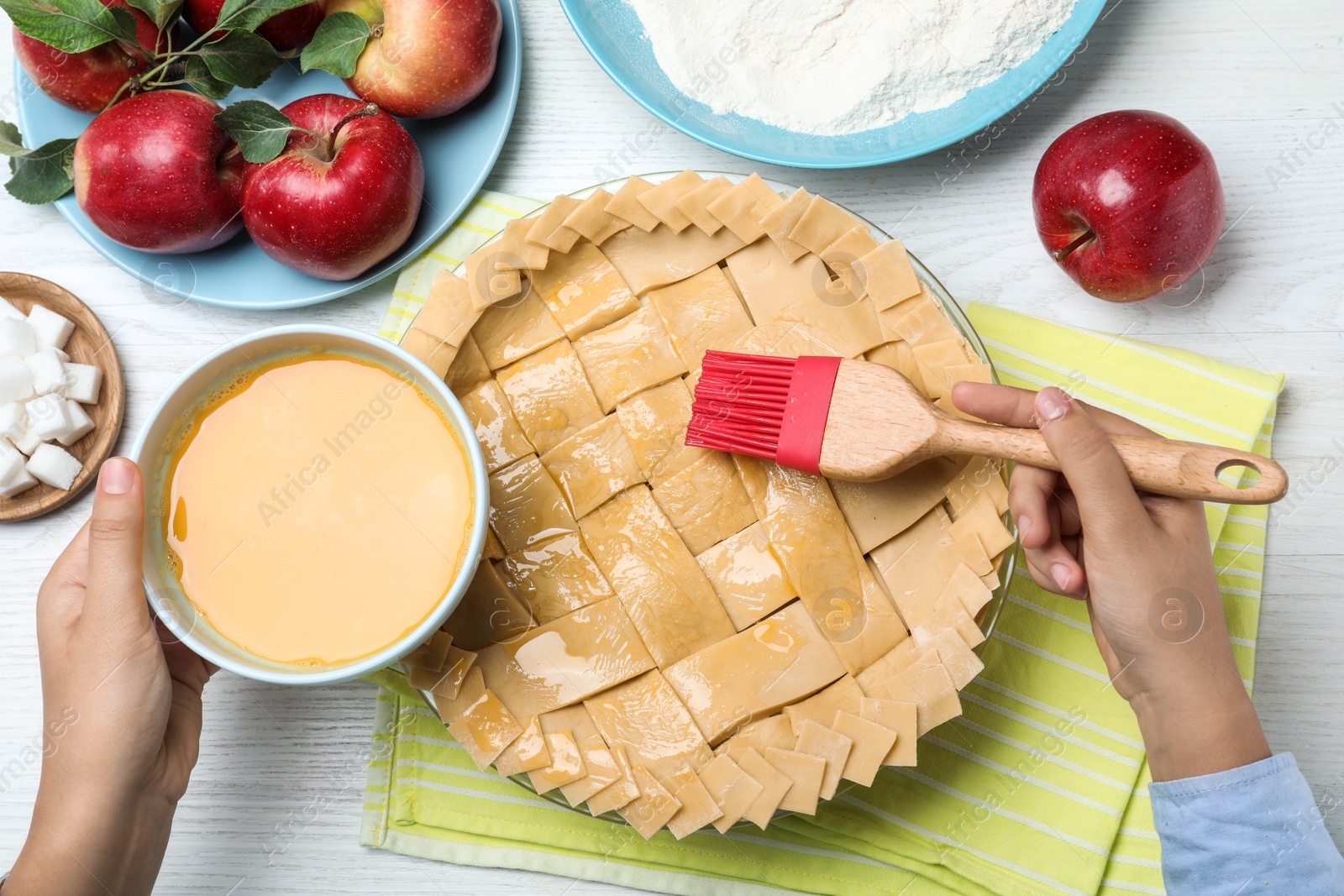 Photo of Woman spreading egg yolk onto raw apple pie at white wooden table, top view