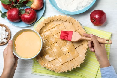 Woman spreading egg yolk onto raw apple pie at white wooden table, top view