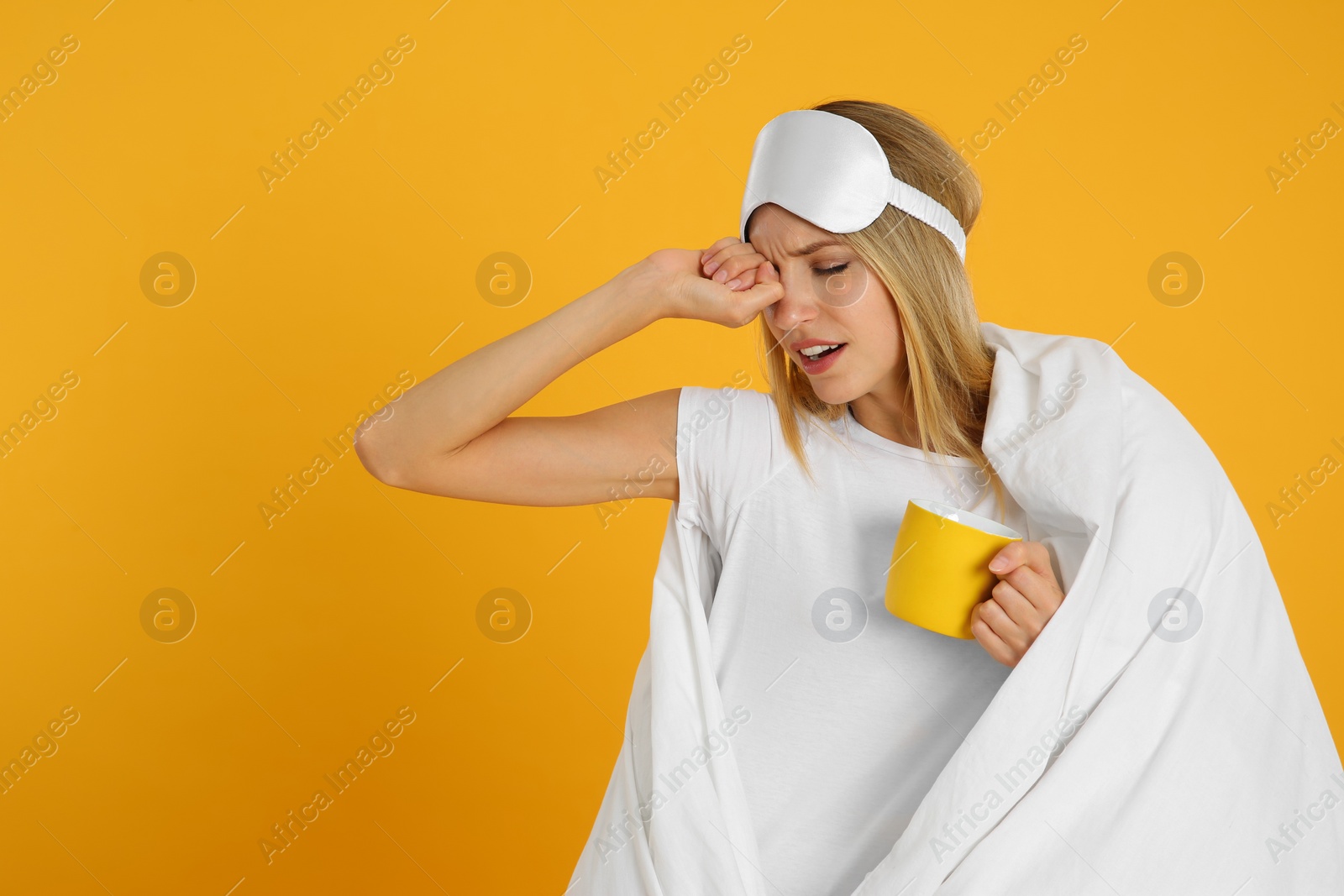 Photo of Young woman in sleeping mask wrapped with blanket holding cup on yellow background