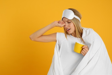 Photo of Young woman in sleeping mask wrapped with blanket holding cup on yellow background