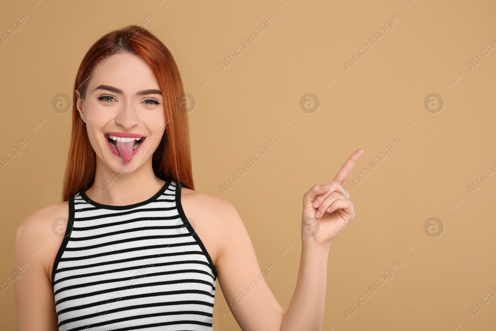 Photo of Happy woman showing her tongue and pointing at something on beige background. Space for text
