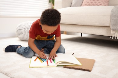 Photo of Cute African-American boy drawing in sketchbook with colorful markers on floor at home