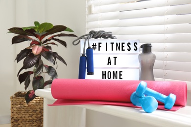 Photo of Sport equipment and lightbox with hashtag FITNESS AT HOME on window sill indoors. Message to promote self-isolation during COVID‑19 pandemic