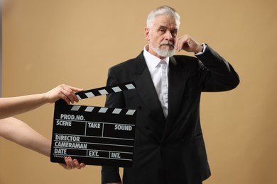 Photo of Senior actor performing role while second assistant camera holding clapperboard on beige background, selective focus. Film industry