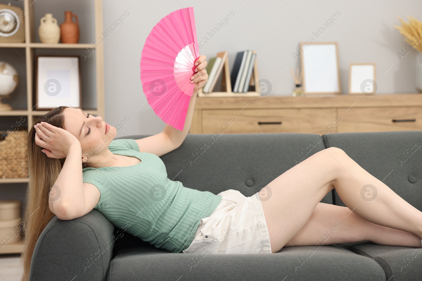 Photo of Woman waving pink hand fan to cool herself on sofa at home