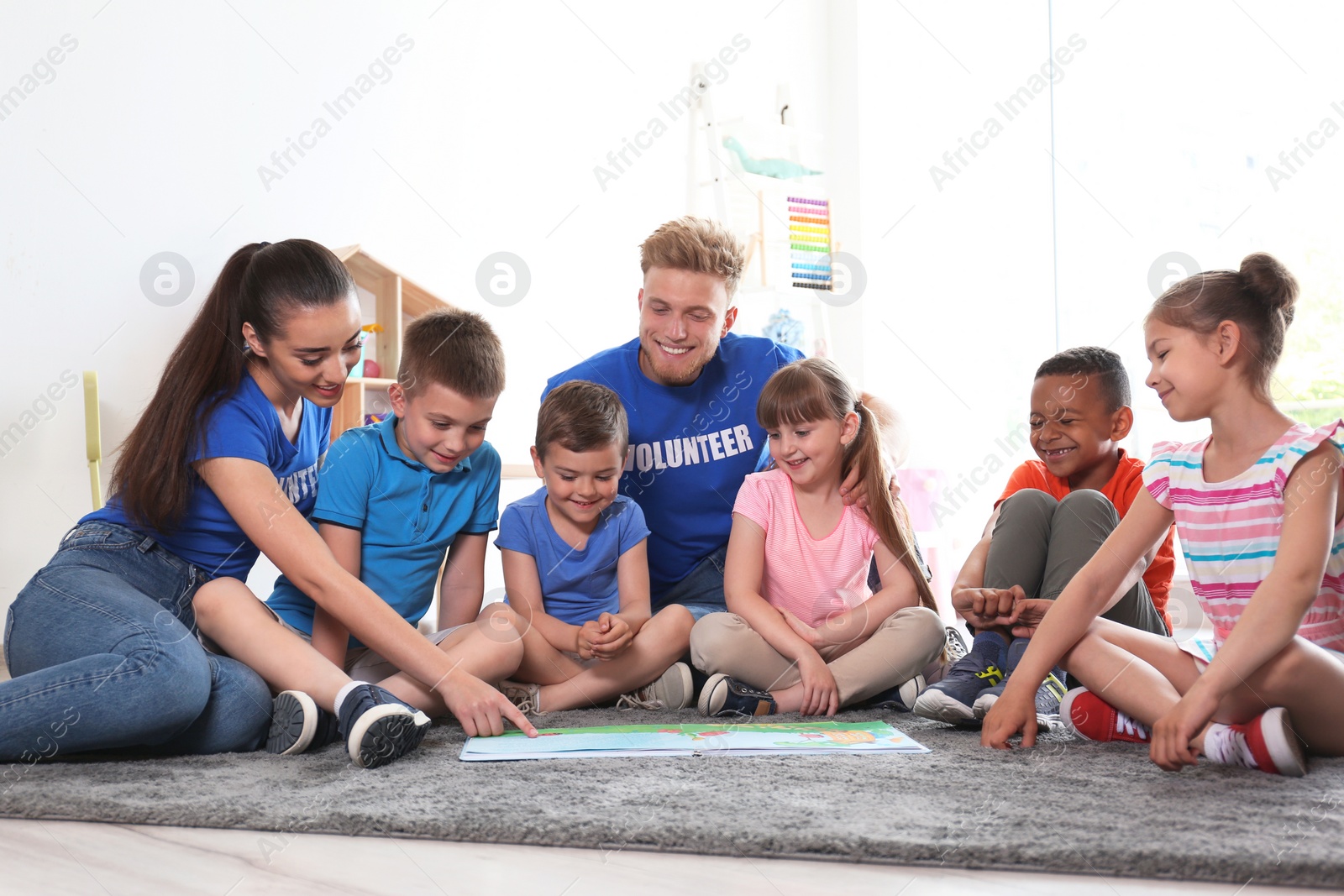 Photo of Young volunteers reading book with children on floor indoors