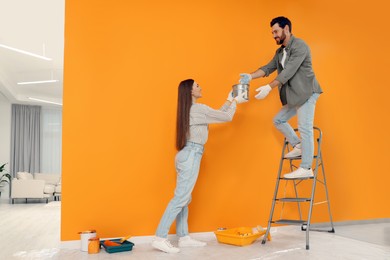 Photo of Woman giving man can of paint near orange wall indoors. Interior design