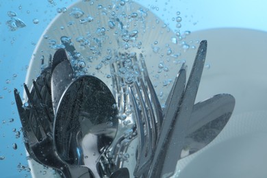 Photo of Washing silver cutlery and plates in water on light blue background, closeup