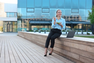 Smiling businesswoman with lunch box sitting near laptop outdoors. Space for text