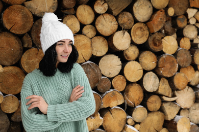 Photo of Young woman wearing warm sweater and hat near stack of firewood, space for text. Winter season