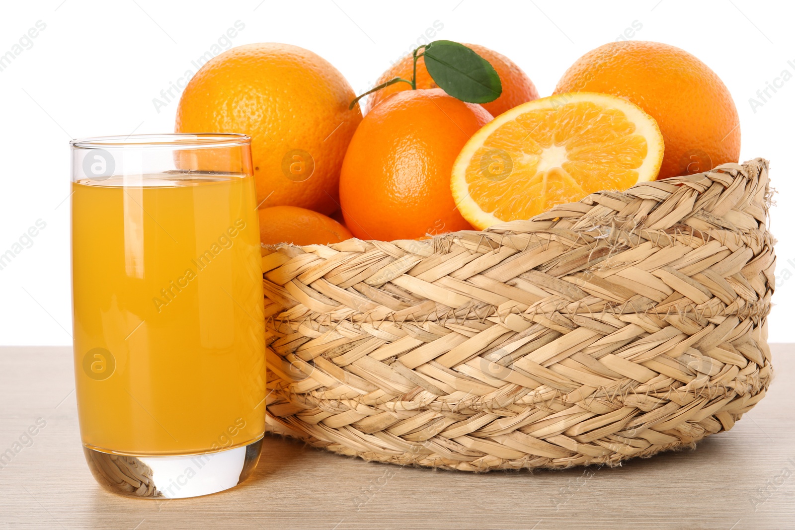Photo of Fresh oranges in wicker basket and glass of juice on light wooden table against white background