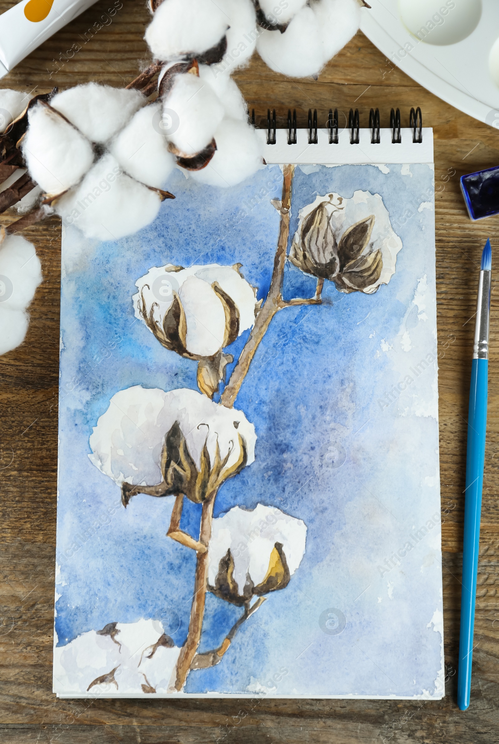 Photo of Painting of cotton flowers in sketchbook and art supplies on wooden table, flat lay