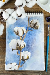 Photo of Painting of cotton flowers in sketchbook and art supplies on wooden table, flat lay