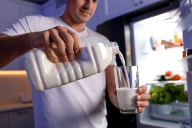 Man pouring milk from gallon bottle into glass near refrigerator in kitchen at night, closeup