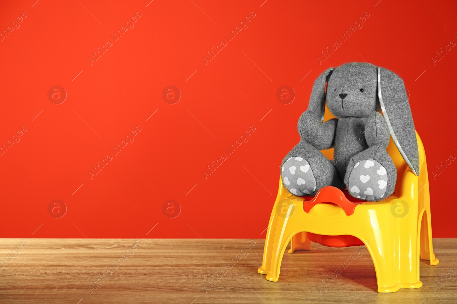 Photo of Grey bunny with yellow potty on wooden table against red background, space for text. Toilet training