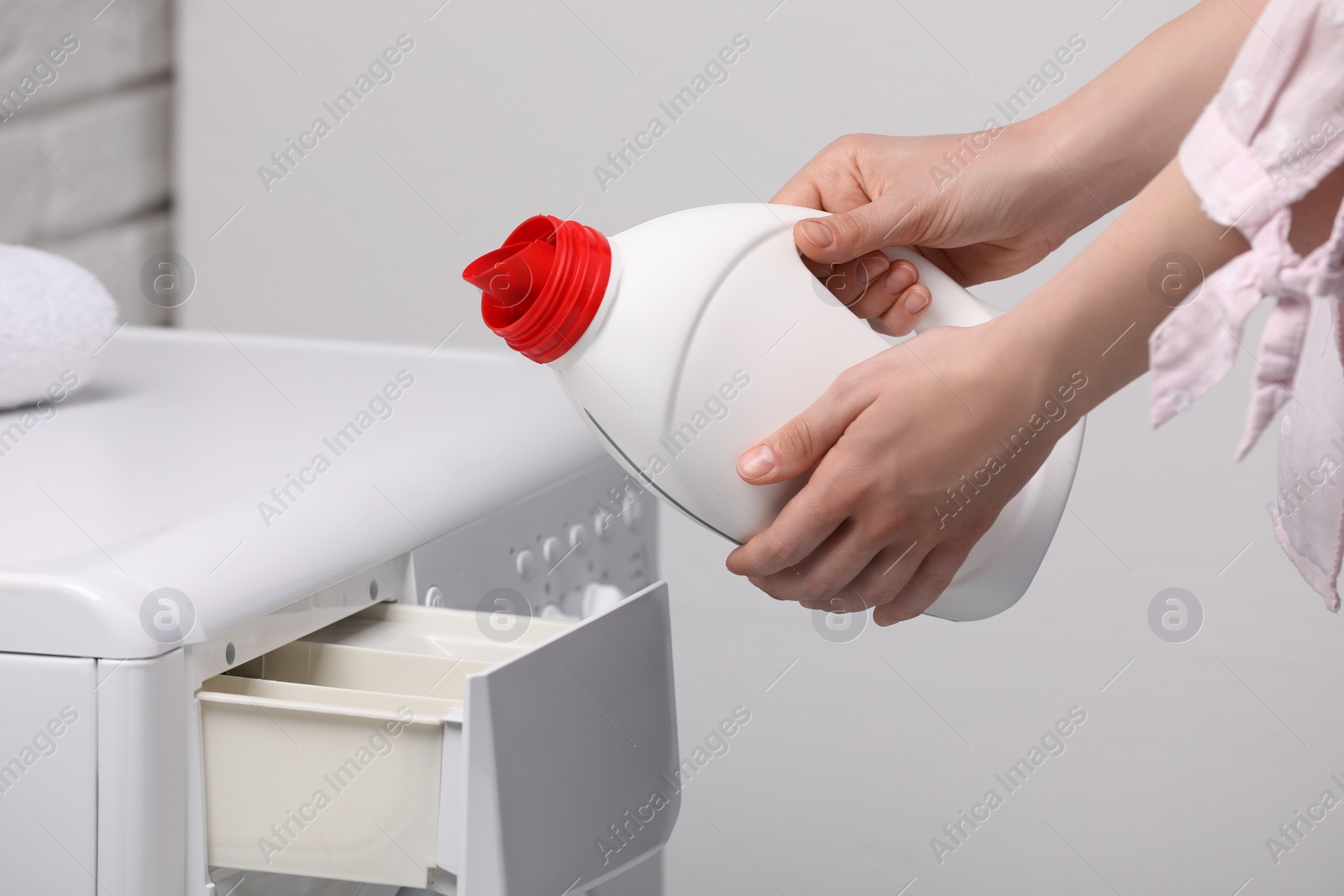 Photo of Woman pouring fabric softener from bottle into washing machine near light grey wall, closeup