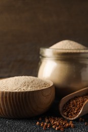 Photo of Bowl of buckwheat flour and scoop with grains on black table against wooden wall, closeup