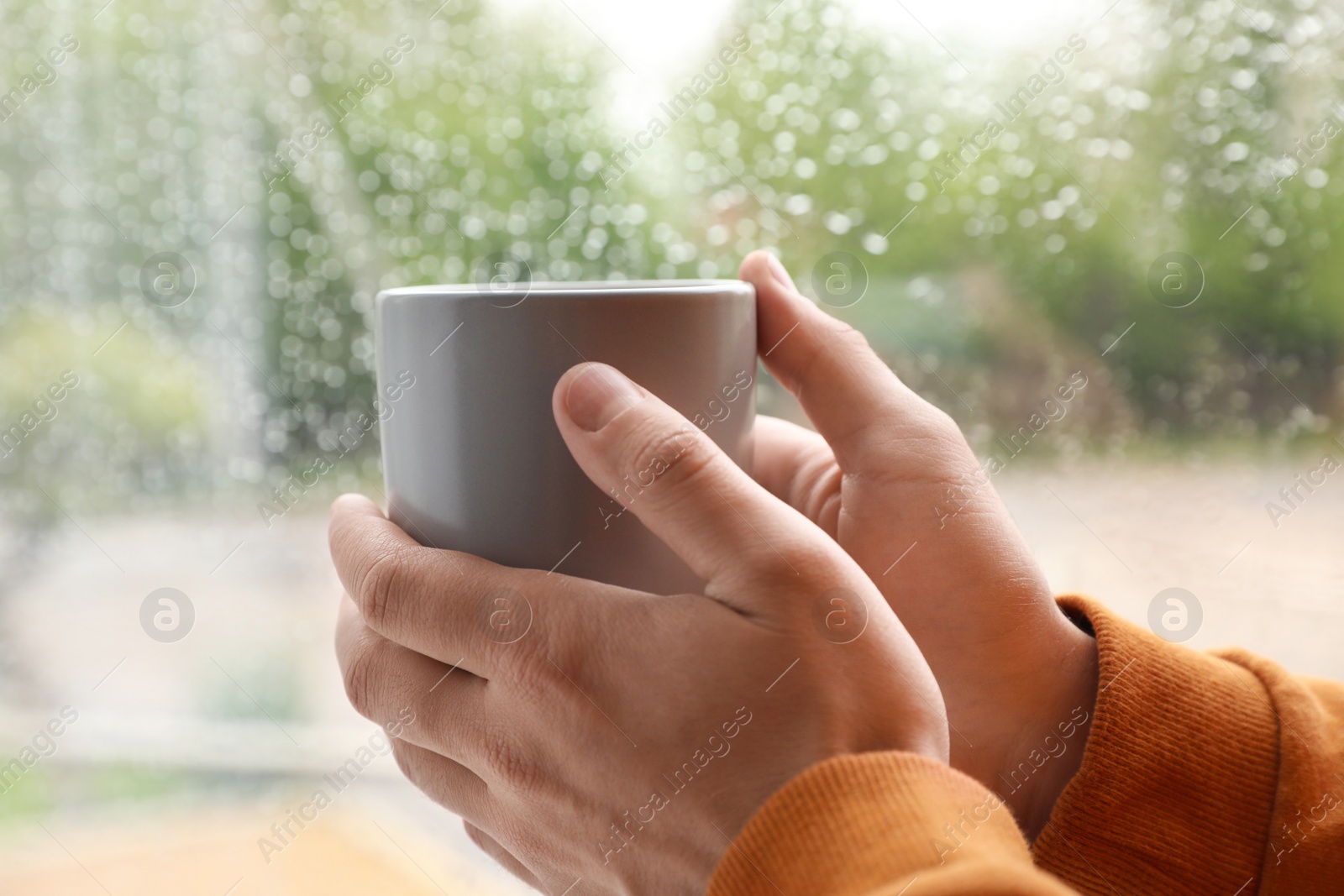 Photo of Young man with cup of coffee near window indoors on rainy day, closeup