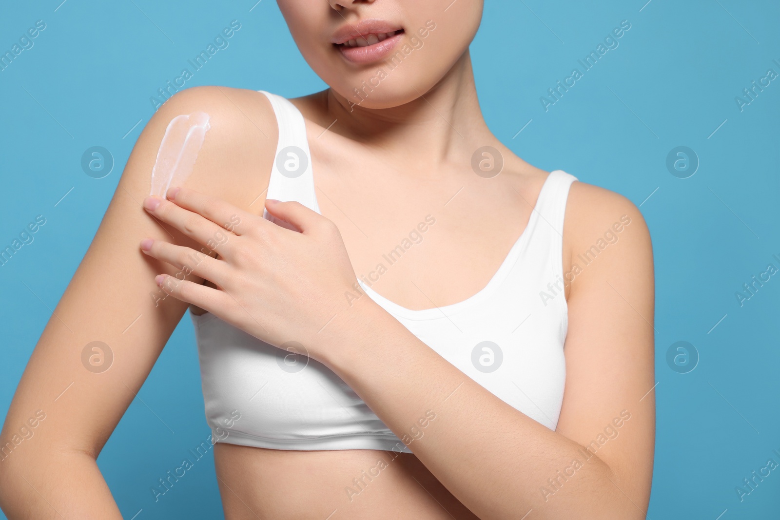 Photo of Young woman applying body cream onto shoulder on light blue background, closeup