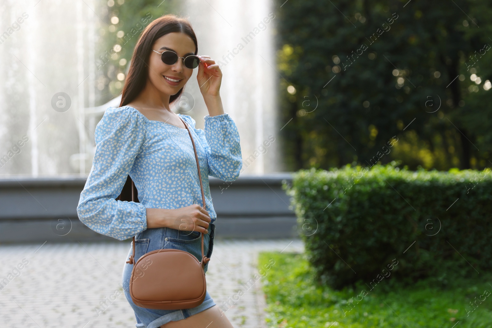 Photo of Beautiful young woman with stylish bag in sunglasses at park outdoors, space for text