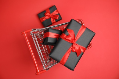 Photo of Small shopping cart with wrapped gift boxes on red background, top view. Black Friday sale