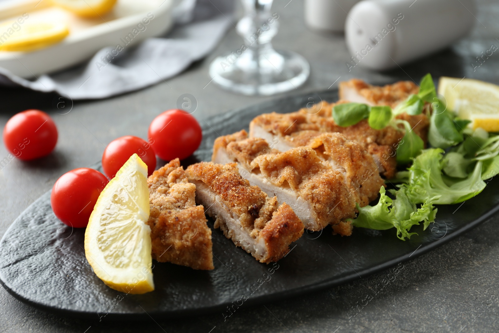 Photo of Tasty cut schnitzel served with tomatoes, greens and lemon on grey table, closeup