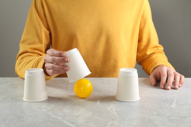 Shell game. Man showing ball under cup at light marble table, closeup