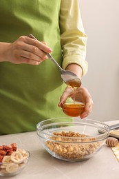Photo of Making granola. Woman adding honey into bowl with mixture of oat flakes and other ingredients at light marble table in kitchen, closeup